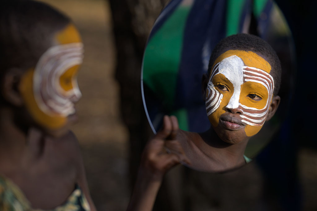 portrait of a young child wearing paint on their face holding a mirror up to themselves